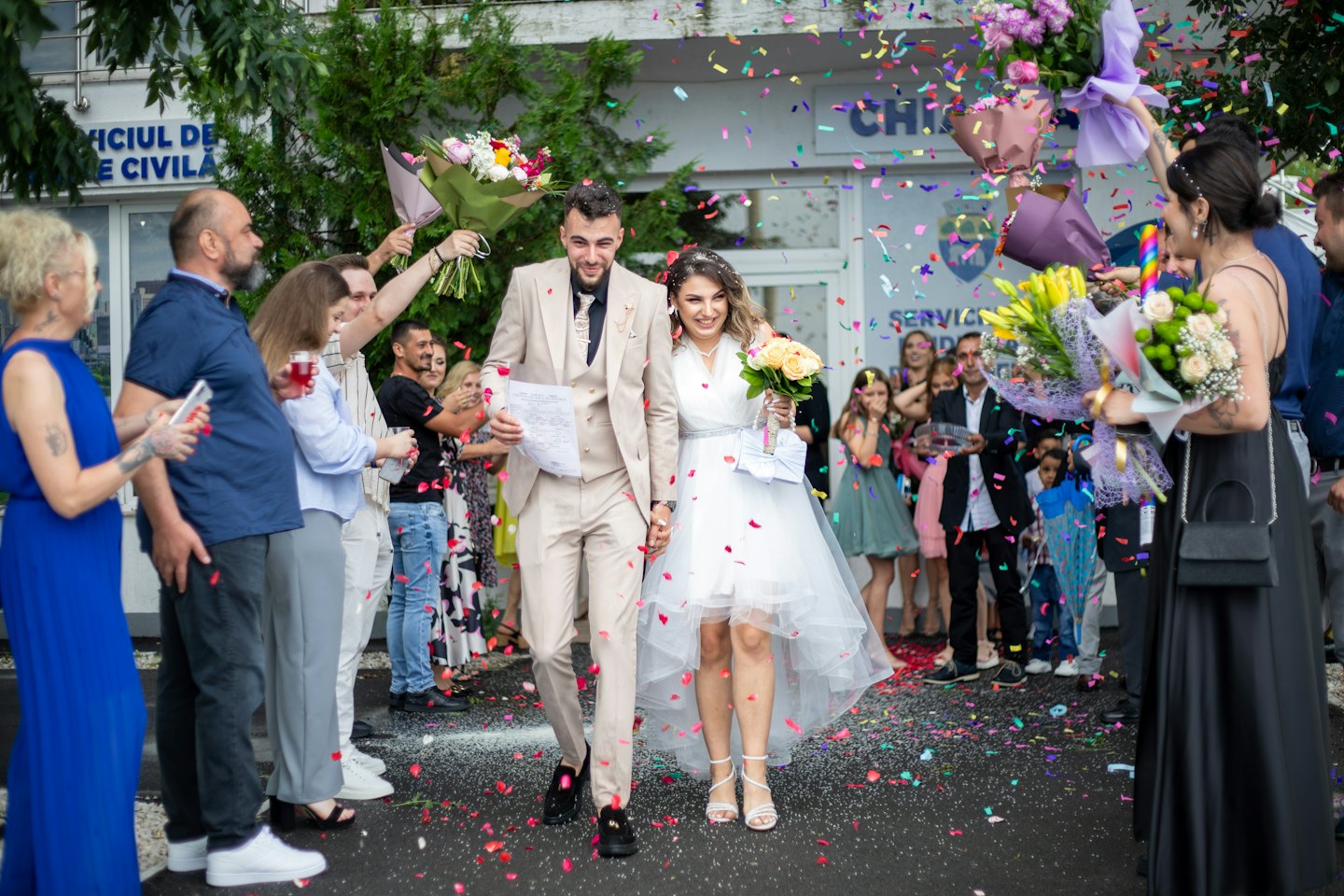 Newlyweds smile as confetti falls outside a building.